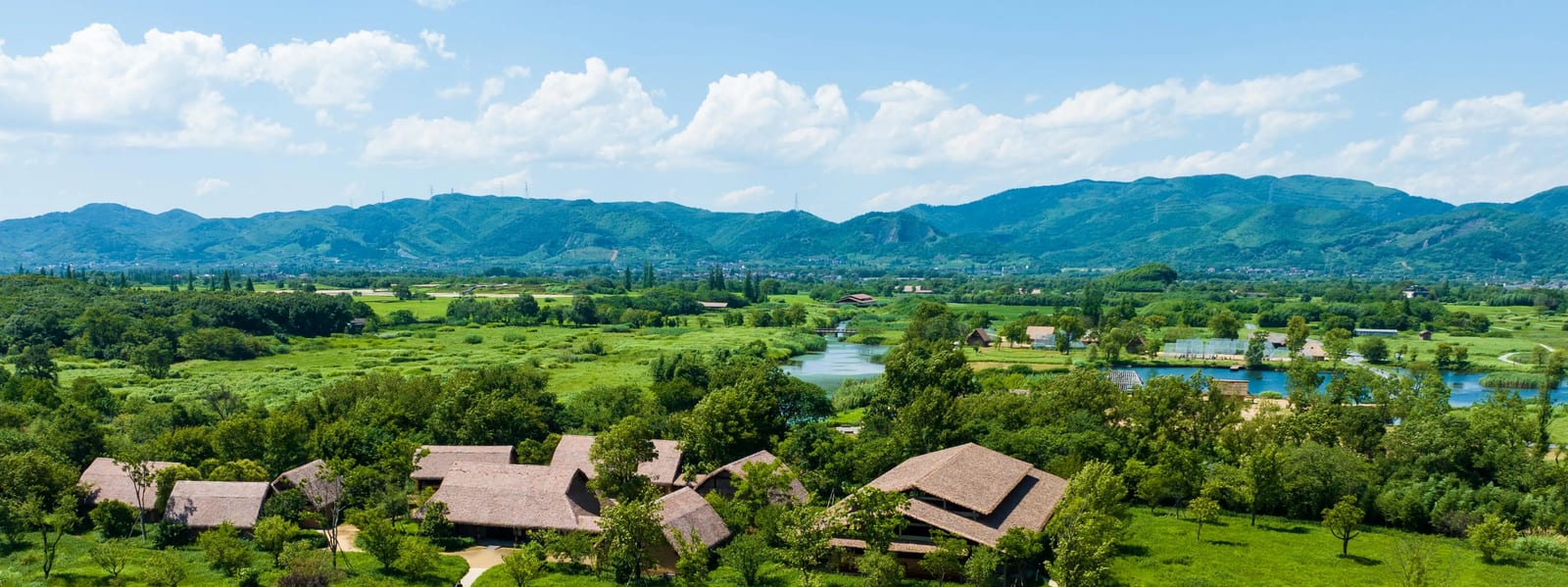 Aerial view across the Liangzhu Ancient City park — reed-thatched buildings in wetland grassland, open water and the hills that ring the floodplain
